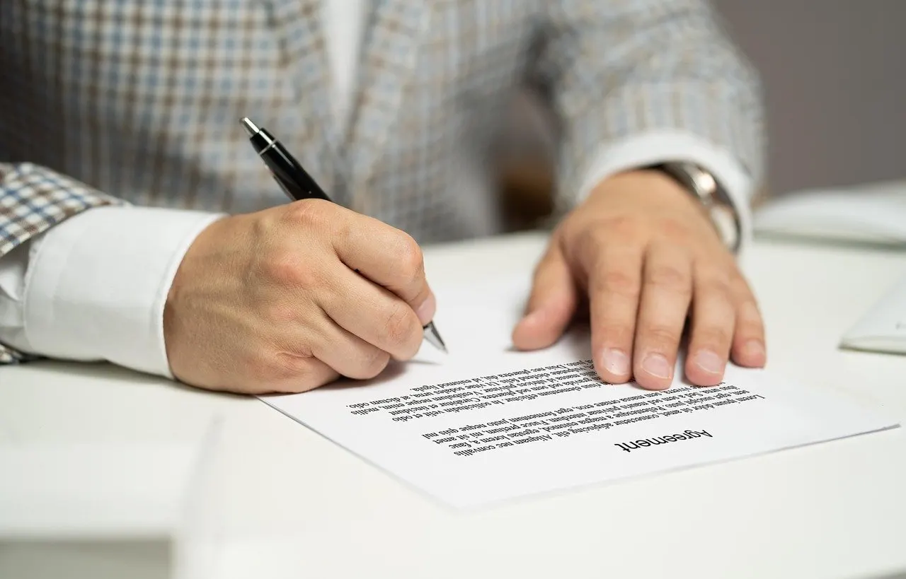 Hand mit schwarzem Kugelschreiber unterschreibt ein bedrucktes Vertragsblatt auf einem weißen Schreibtisch, Ärmel eines karierten Sakkos