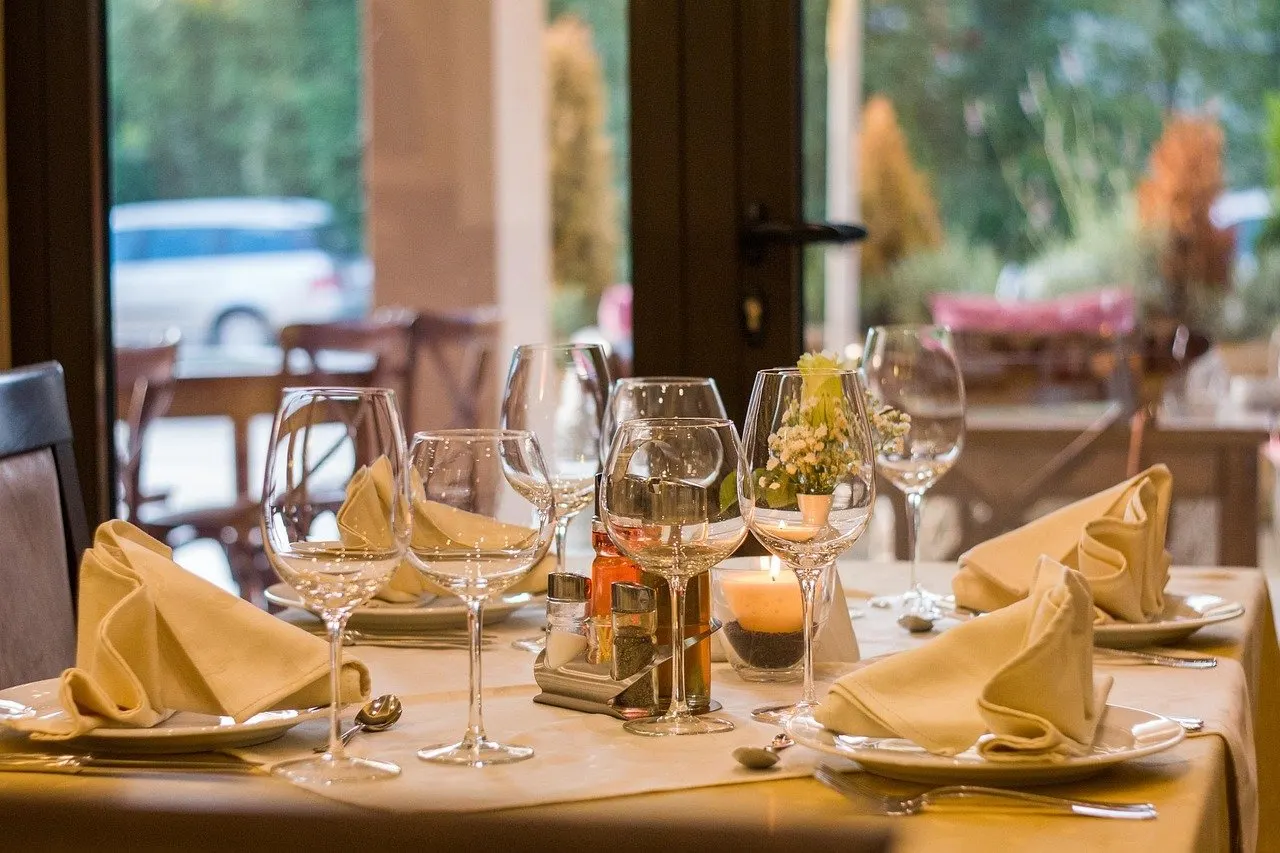 Eingedeckter Restauranttisch mit Weingläsern, gefalteten Stoffservietten, Tellern und Besteck vor einer großen Fensterfront