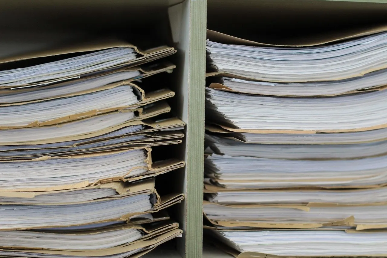 Zwei Regalfächer, dicht gefüllt mit gestapelten Papierakten in hellbraunen Aktendeckeln, die Blätter leicht gewellt