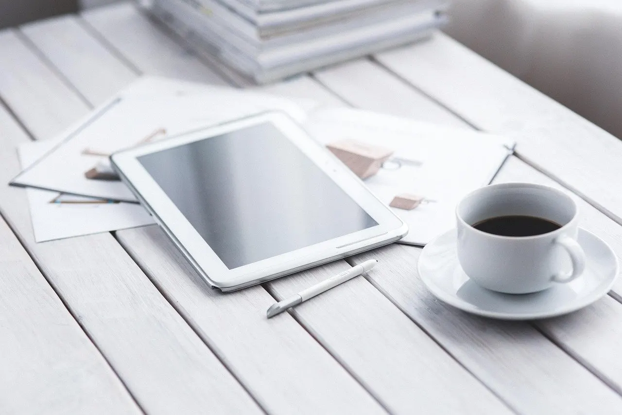 Weißes Tablet mit Eingabestift auf einem weiß lackierten Holztisch, daneben ausgedruckte Unterlagen und eine Tasse schwarzer Kaffee