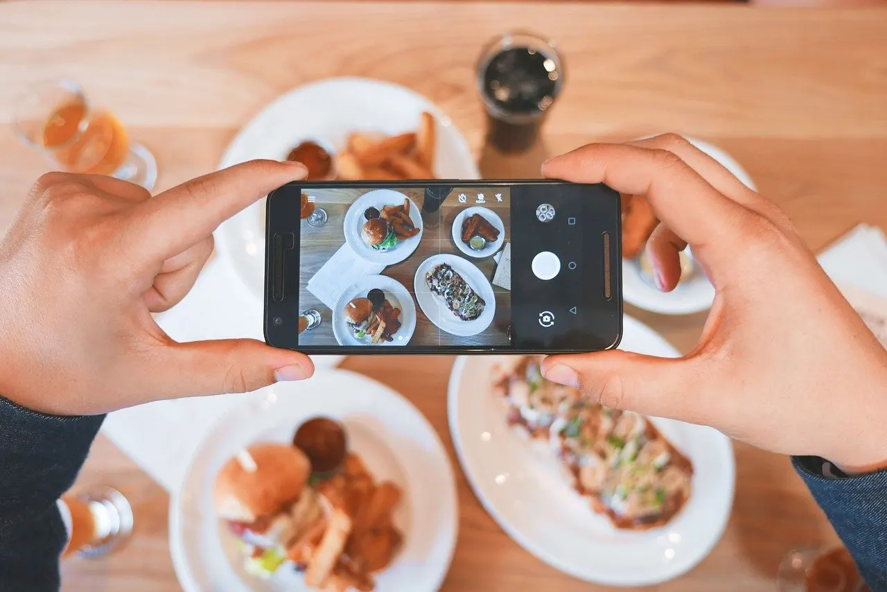 Zwei Hände halten ein Smartphone über einen Restauranttisch und fotografieren Teller mit Burger, Pommes und weiteren Gerichten