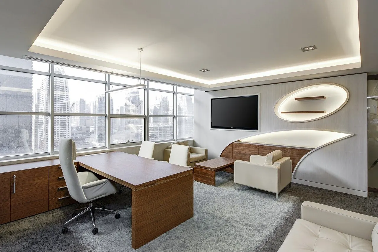 Chefbüro mit Holzschreibtisch, weißem Bürodrehstuhl, hellen Polstersesseln, Wandfernseher und großer Fensterfront zur Stadt