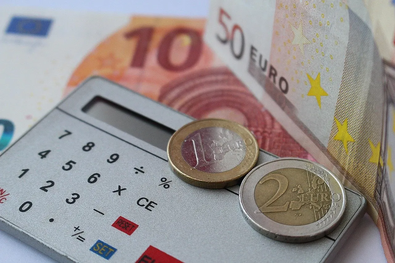 Grauer Taschenrechner mit einer Ein- und einer Zwei-Euro-Münze darauf, dahinter Zehn- und Fünfzig-Euro-Scheine