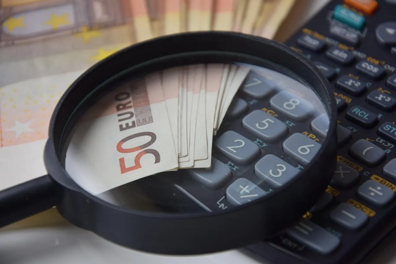 Lupe über einem Fächer aus 50-Euro-Scheinen, daneben ein schwarzer Taschenrechner, weitere Banknoten im unscharfen Hintergrund