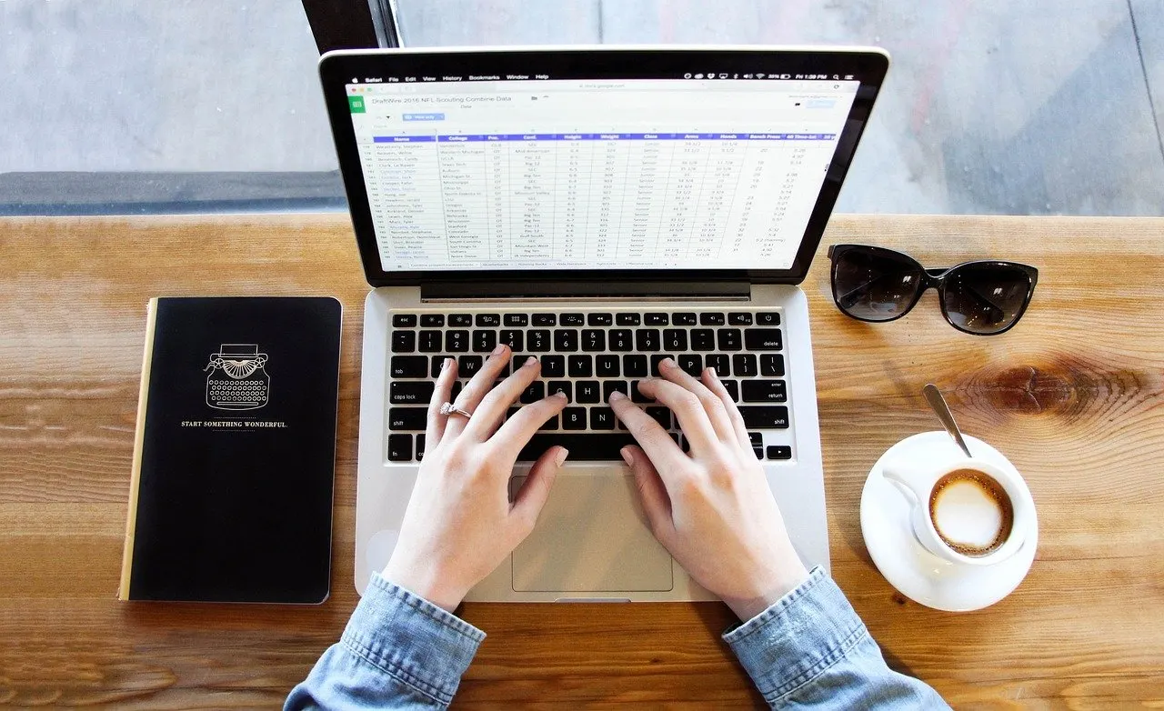 Hände tippen auf einem Laptop mit geöffneter Tabellenkalkulation, daneben Notizbuch, Sonnenbrille und Kaffeetasse auf Holztisch